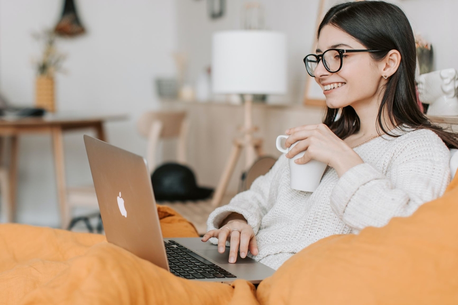 A student sits on a couch with a laptop on their lap and smiles as they look at the screen.